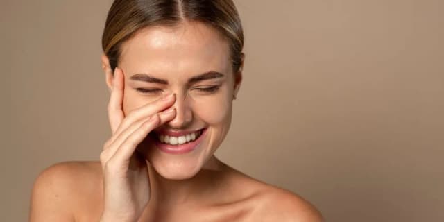 The woman stands against a beige wall and laughs and covers her face with her hand. 